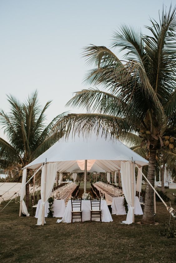 Wedding reception venue featuring an outside tent and long dressed tables with palm trees in background