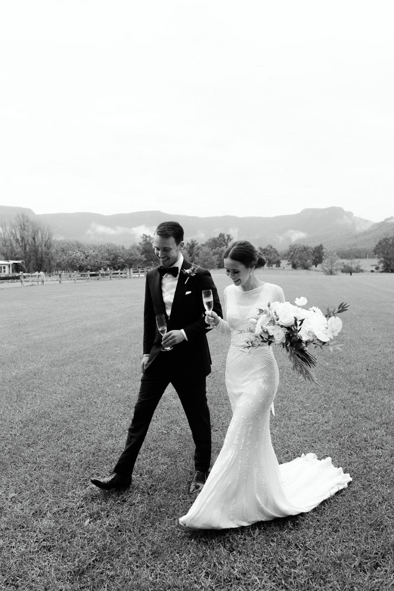 B&W image of Karen Willis Holmes bride Anna walking with husband Julian. Holding her bridal bouquet and glass of champagne. Wearing the Cassie wedding dress.