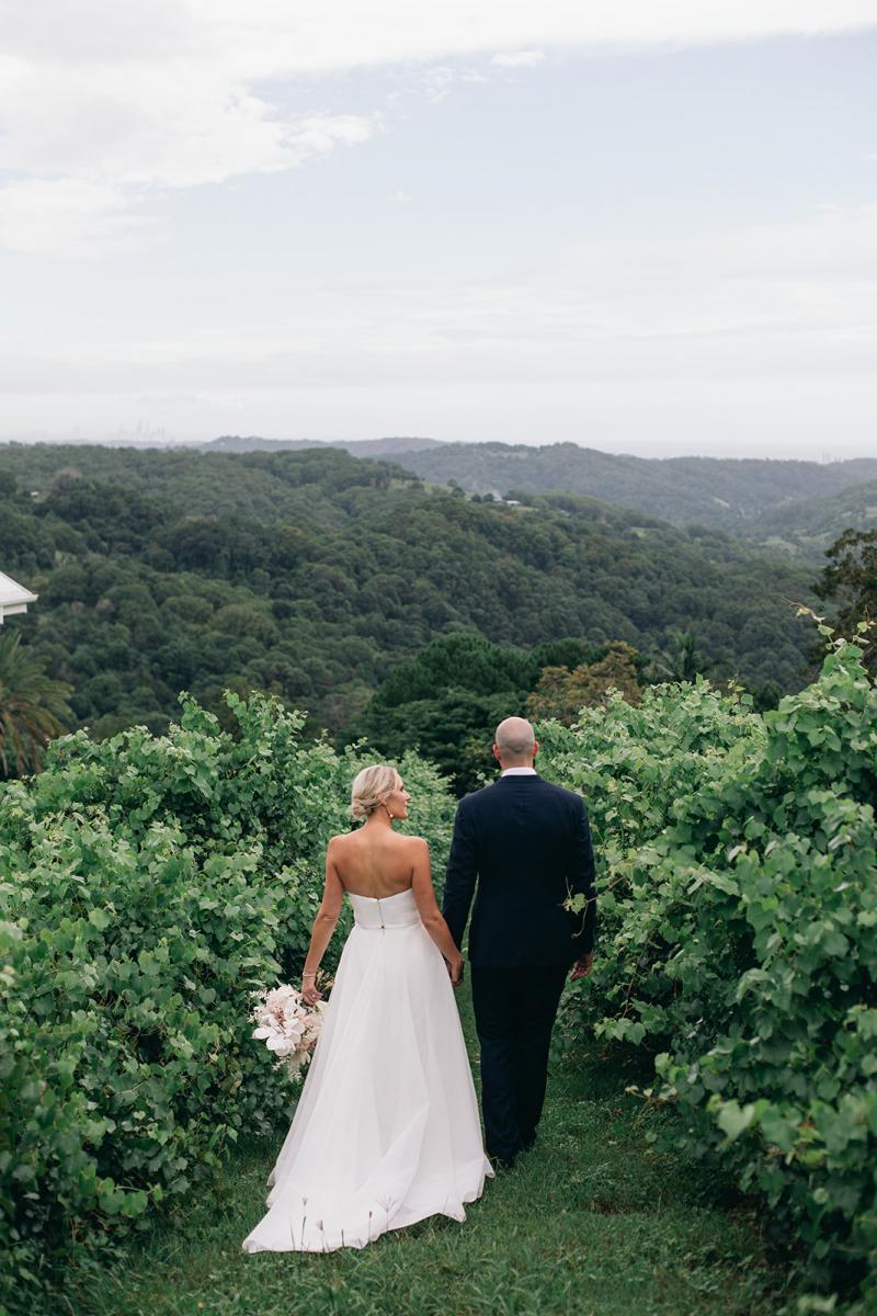 KWH bride Samantha and husband Adam walking through a vineyard. Samantha holds a timeless bouquet and wears the Esther wedding dress with a long train made of mesh..
