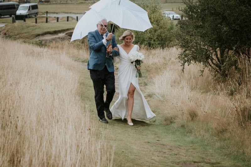 Real bride Lauren and her dad walk down the grass aisle in Queenstown wedding ceremony. Father holds the umbrella over both as Lauren's Nikki gown from KWH, splits open as she walks.