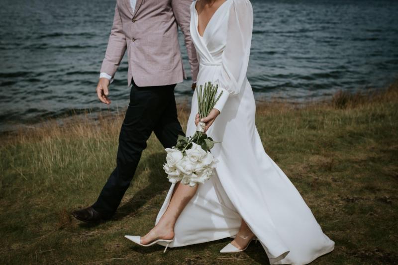 Real bride Lauren walks with new husband, Will, by the lake side in Queenstown after their small wedding ceremony. Lauren wears the modern NIkki wedding dress.