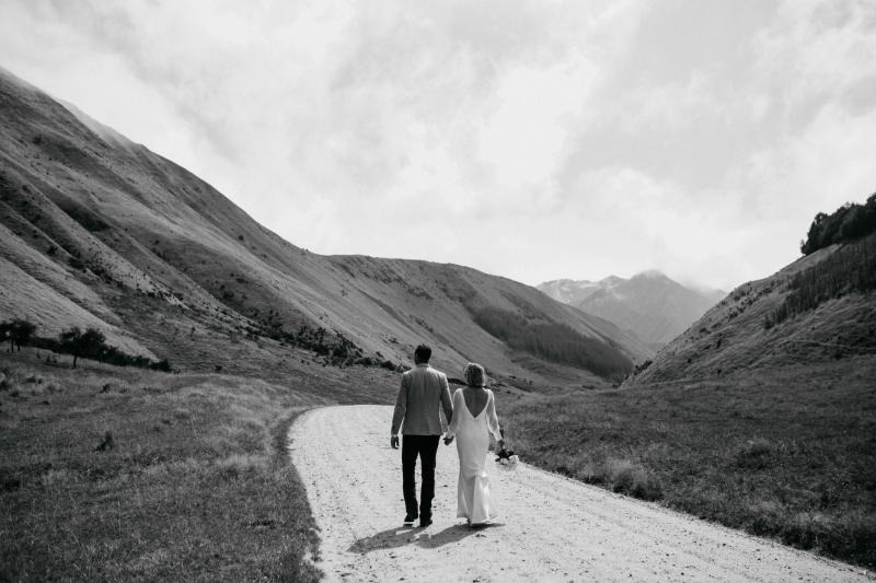 New married couple, Will and Lauren, walk away on a winding path in the New Zealand hills, while she wears the simple Nikki gown with low back by KWH.
