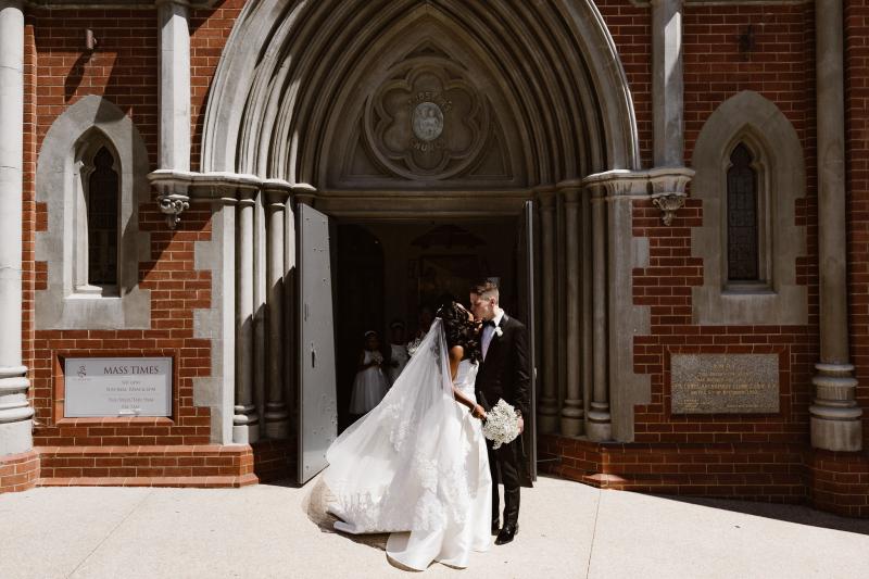 Real bride Ancille kisses her new husband under the church's arched doorway. The bride wears the minimalist Kitty Melanie gown from KWH.