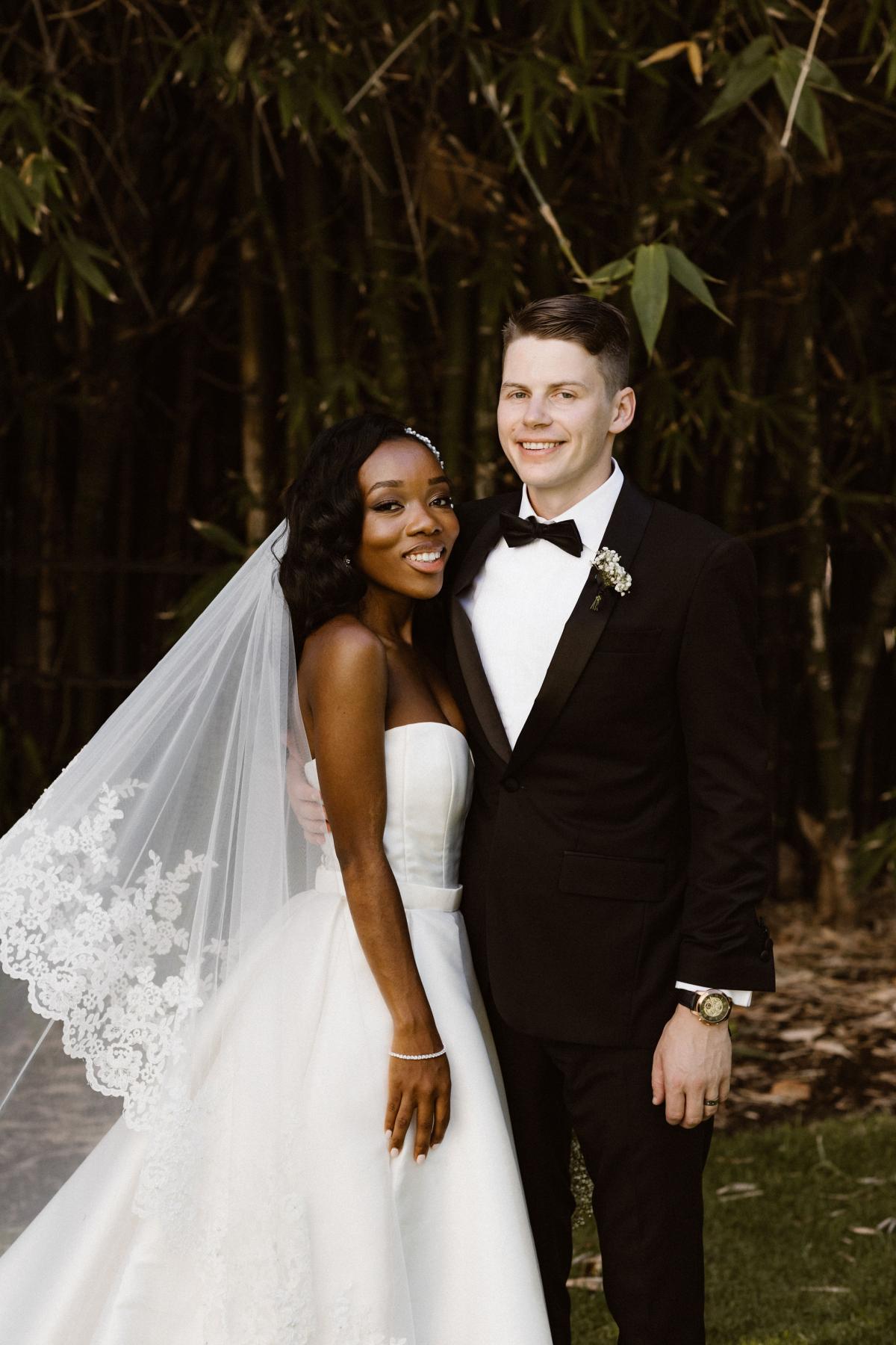 Real bride Ancille stands with new hubby Dar in front of foliage. She pairs her classic Kitty Melanie gown by KWH with a detailed lace veil by Steven Khalil