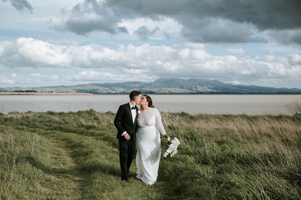 KWH bride, Jarna, stuns in gorgeous CURVE Karina modern lace dress, with long sleeves, and illusion neckline as she kisses her new husband in front of a lush, mountainous New Zealand backdrop.