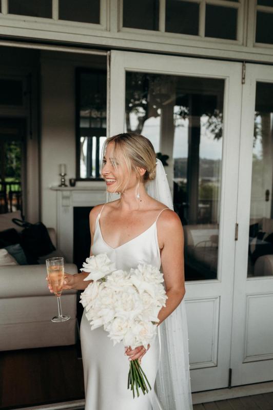 Riley stands in the doorway with mimosa and bouquet in hand as she gets ready to walk down the aise in her Sage gown by Karen Willis Holmes; she features a deep v-neck and pluding back.