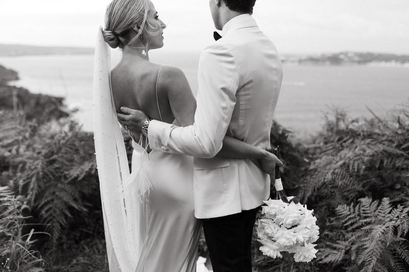 B&W picture of KWH real bride Riley and Wes side hugging in front of ocean scenery. The bride wears the minimalist Sage gown with plunging back and feather tastles.