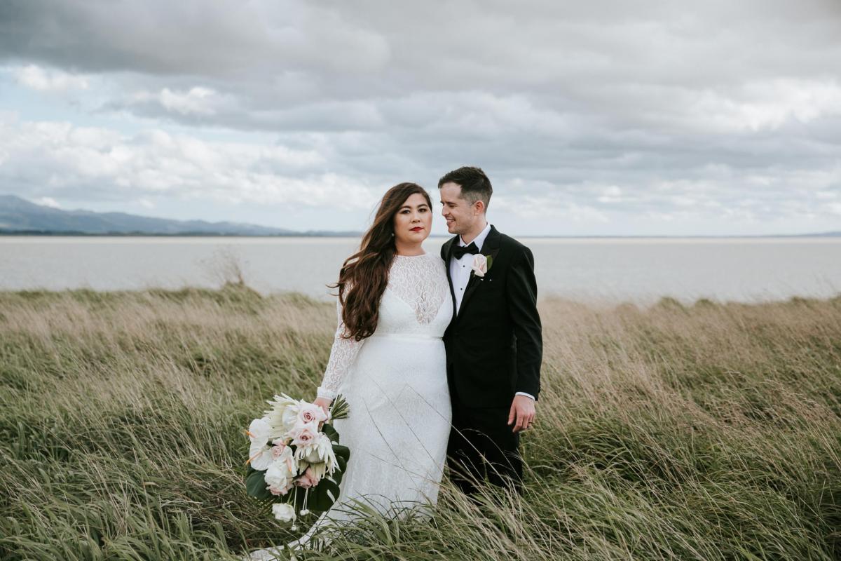 Real bride Jarna stares into the lens as husband Aden loving looks at her as they stand in the stunning New Zealand countryside for their timeless wedding.