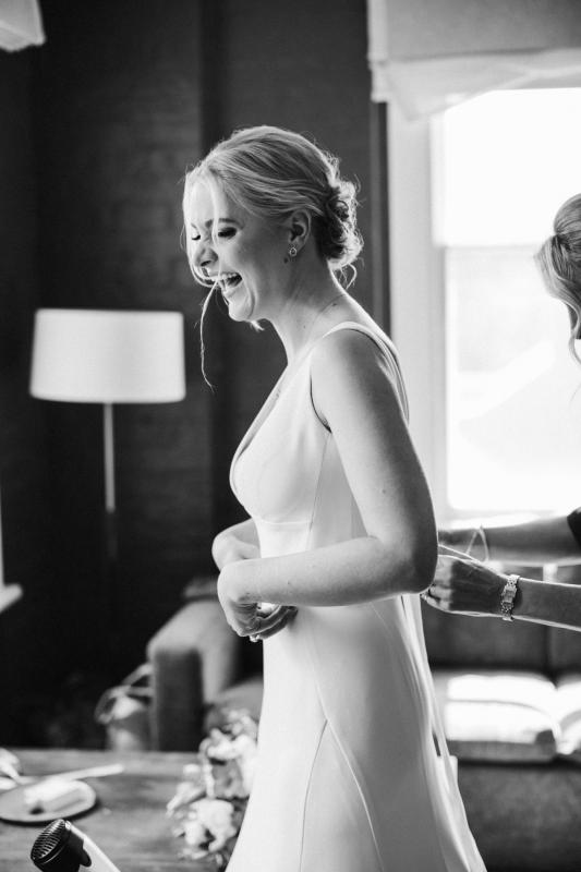 Real bride Eliza laughing as she gets ready in this B&W photo. She wears the modern but elegant Imogen gown from KWH.