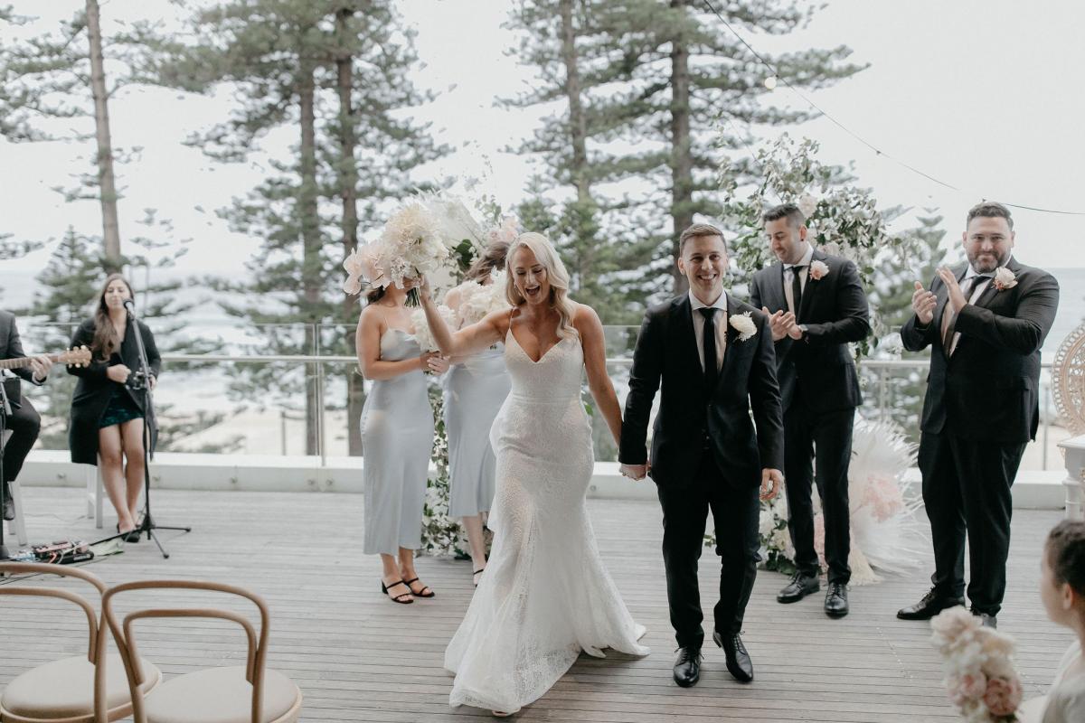 Real bride Nicole and Chris walking downt the aisle holding hands. She wears the minimalist lace Justine gown by KWH with detachable Odette train.