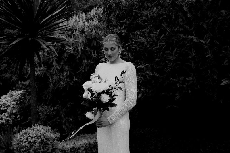 Real bride Hannah standing with her white bridal bouquet while wearing the beaded Margareta gown by KWH in this black and white photograph.