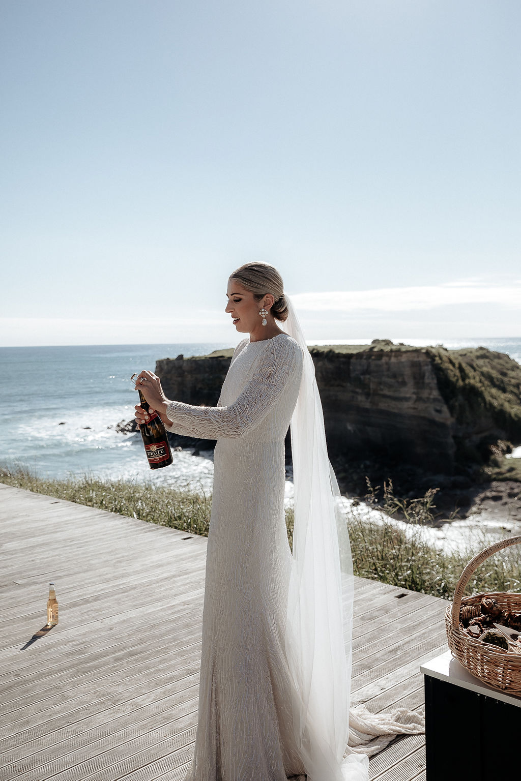 KWH real bride Hannah pops a bottle of bubbly with the New Zealand coast in the background. She dons the trendy sequined Margareta gown with low back and long sleeves.