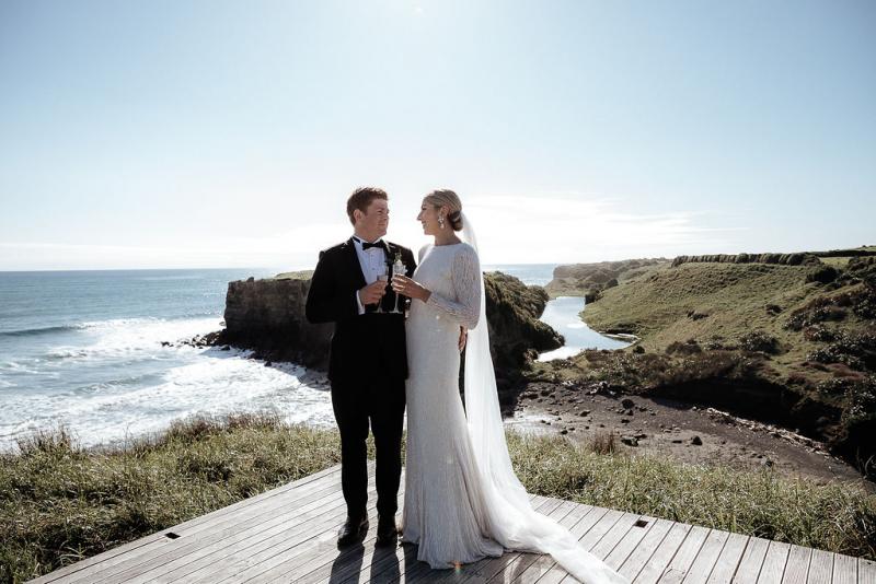 KWH real bride Hannah and Angus stand at the shoreline with drink in hand for a toast. She wears the modern beaded Margareta wedding dress with plunging back.