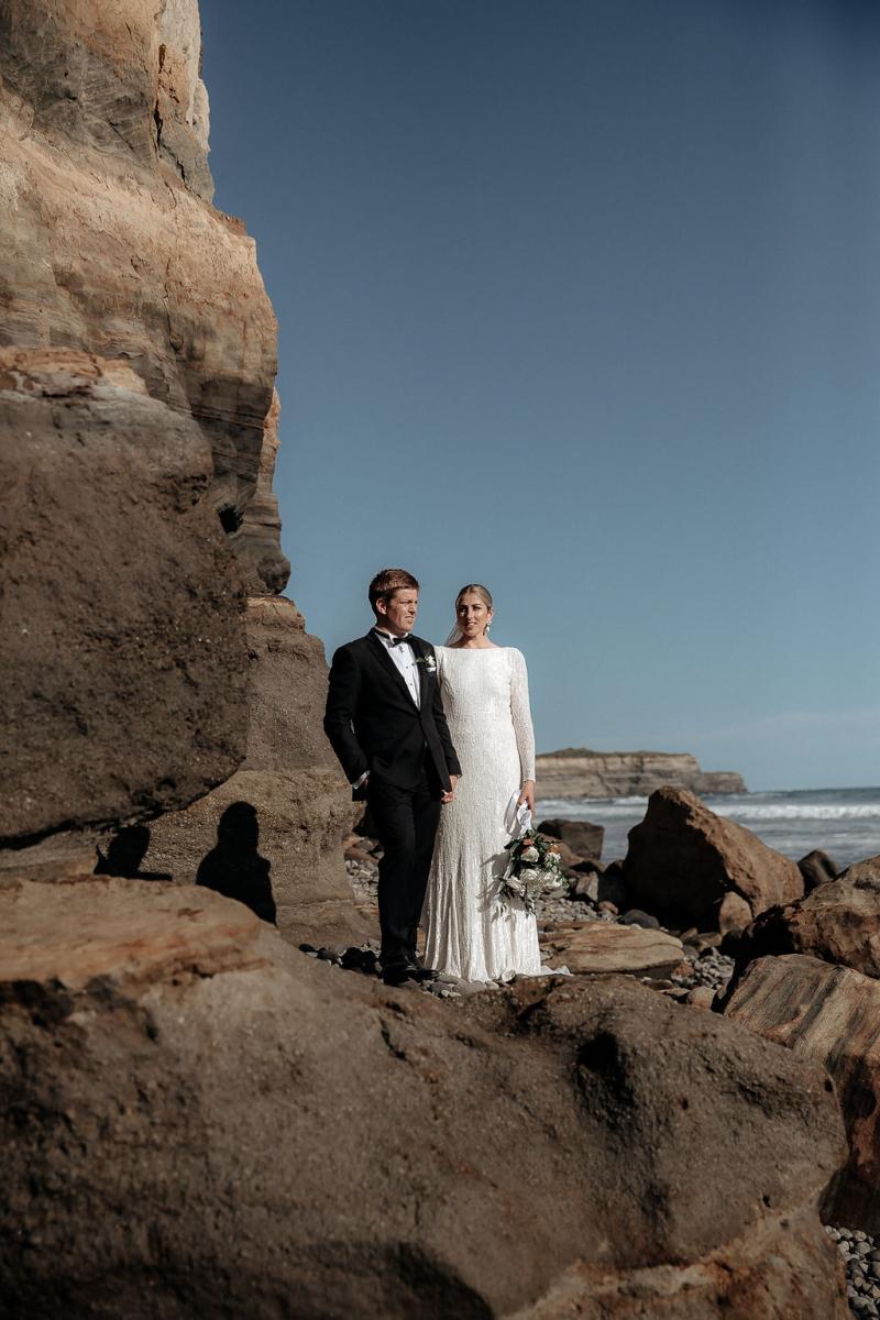 Karen Willis Holmes Real Bride Hannah and Angus stand on a stone formation on the beach. She wears the effortless Margareta gown.