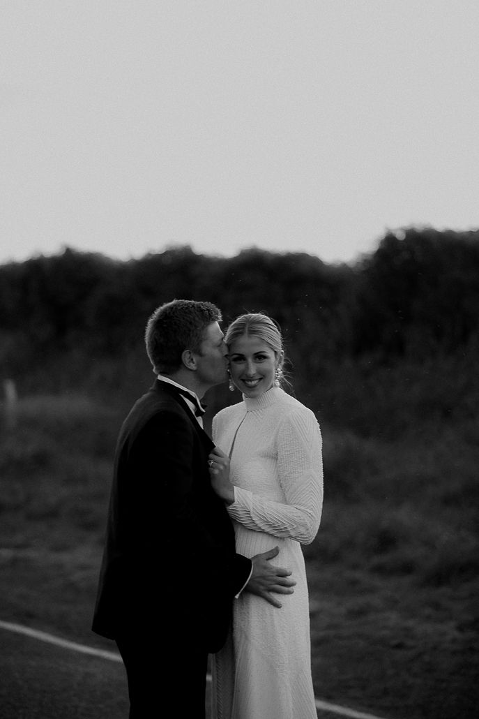 Black and white image of Real Bride Hannah and Angus standing together. She wears a custom designed gown made by her brother in NYC.