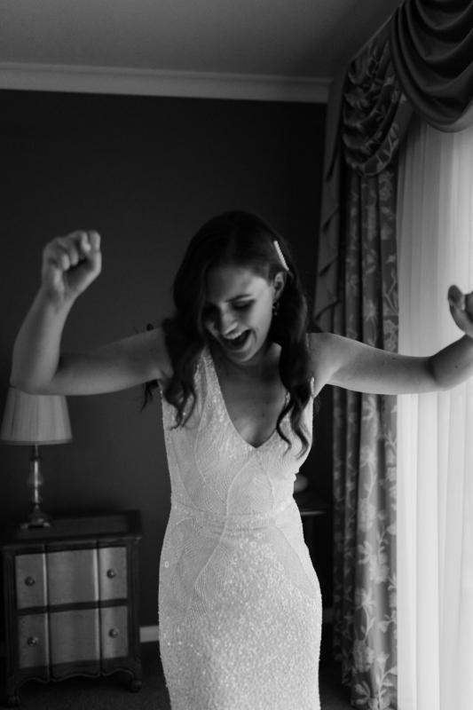 Black and white photo of Real Bride Amber dancing in the room, getting ready for the big day. She wears the v-neck Olympia gown from KWH.