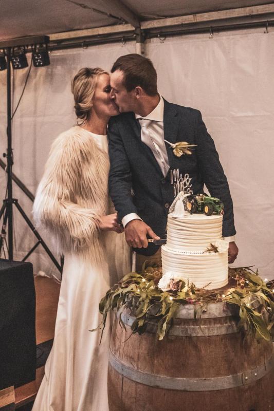 KWH real bride Angela kissing Graham while cutting their country inspired wedding cake. She wears a fur cape over her simple Bridget gown.
