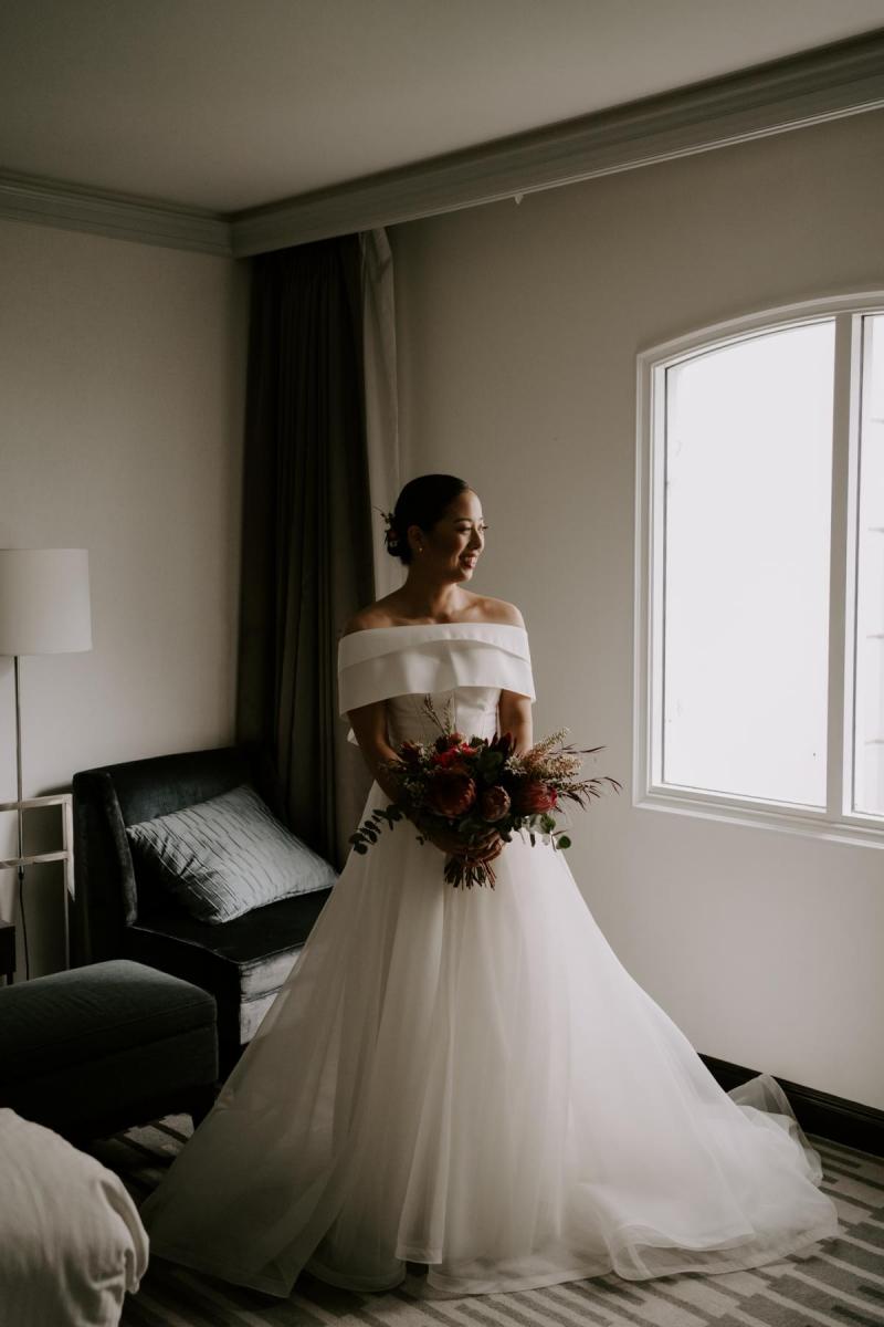 KWH real bride Sarah in front of a window getting ready in her Kitty Joni gown with a-line tulle skirt.