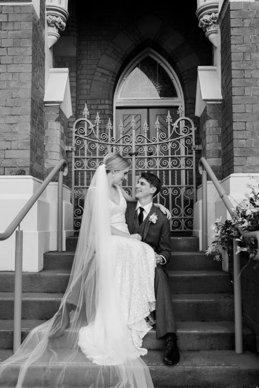 KWH real bride Katie sits on Adrian's lap on the curch steps. She wears the classy sequined Lola gown with fit and flare silhouette.