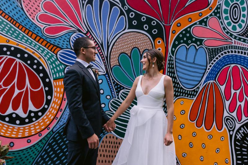 KWH real bride Annalese stands with Hilton in front of a vibrant mural. She wears the ivory Aisha gown with rosette and aline skirt