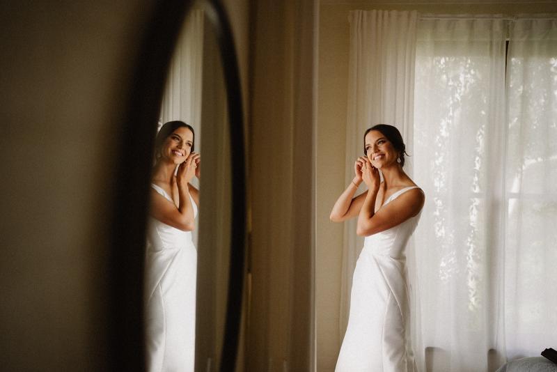 Real bride Charlotte getting ready in the mirror in her Leonie Alexia gown by KWH which features a U- neck and fitted skirt.