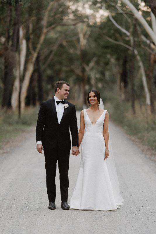 Real bride Charlotte and Myles stand in the middle of a tree lined street together. She wears the ivory Leonie Alexia gown by KWH.