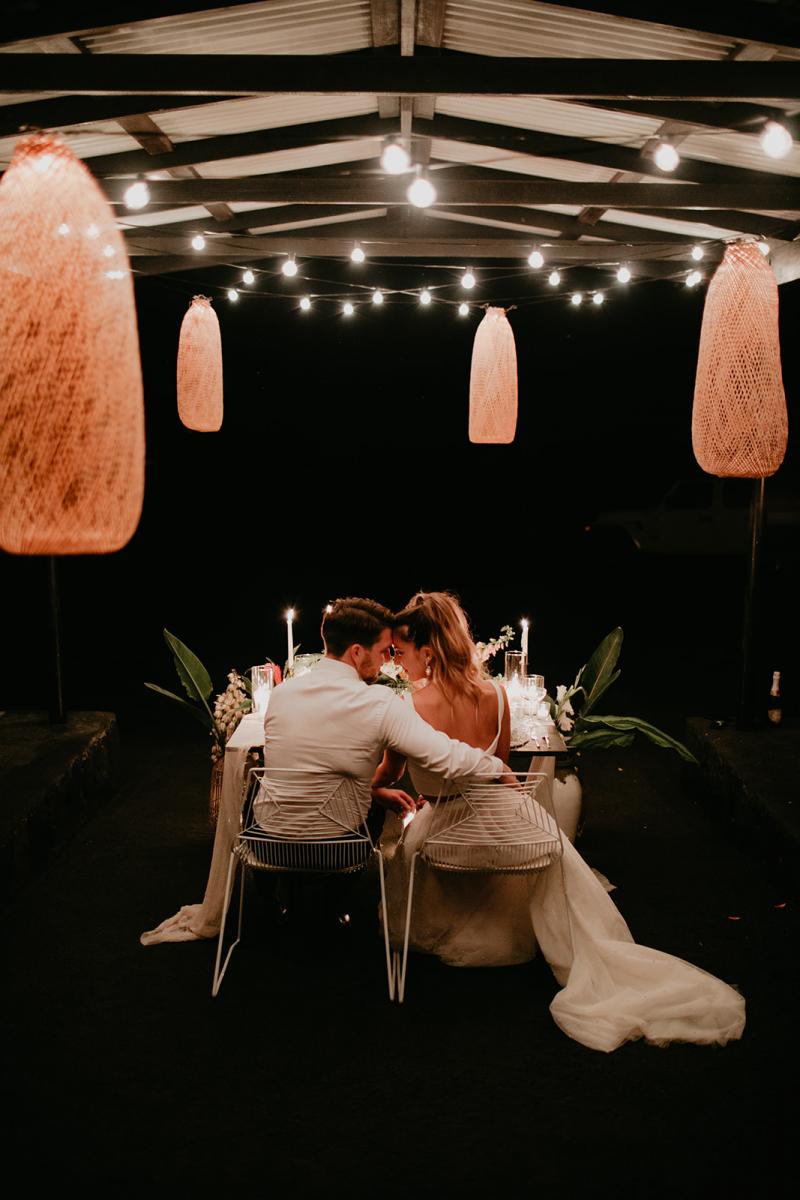 KWH real bride Jana and Garret sitting at their wedding table under the string lights. She wears the voluminous Erin and Lea two piece wedding dress.
