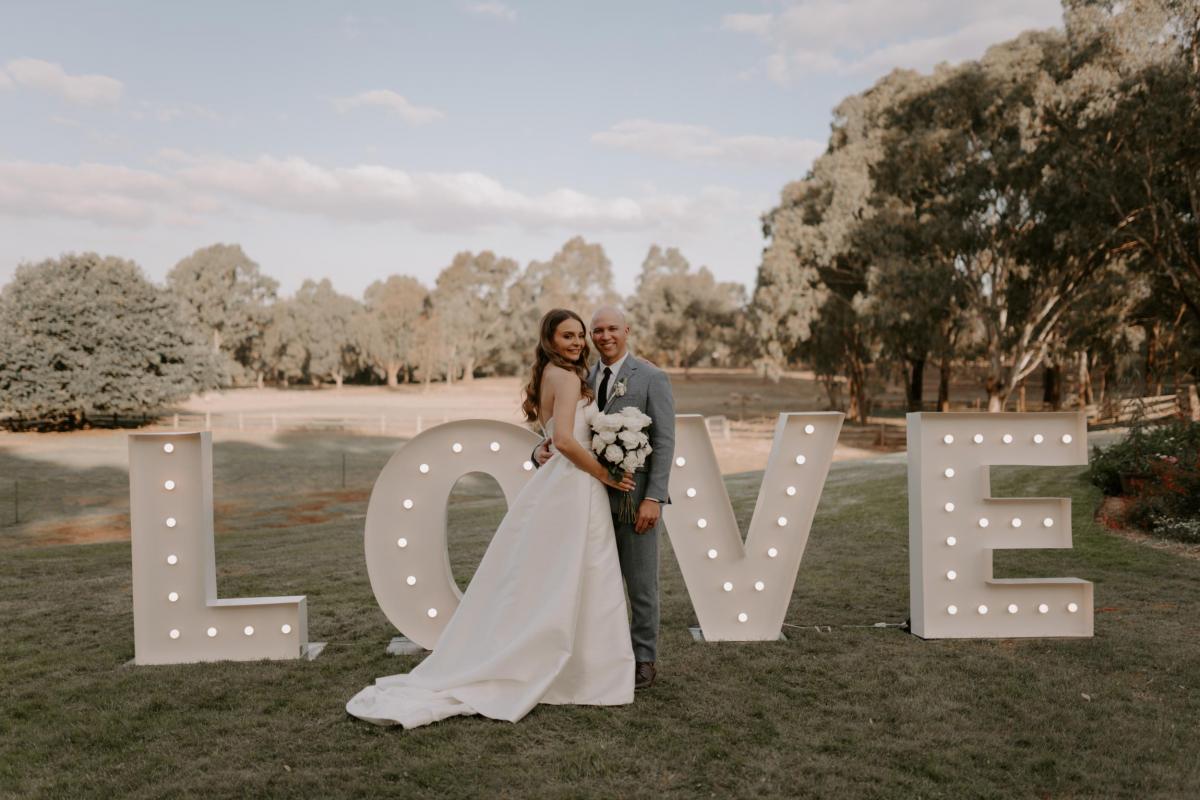 KWH real bride Elke and husband Kyle stand in front of large "love" letters. She wears the classic Blake Camille wedding dress with pockets