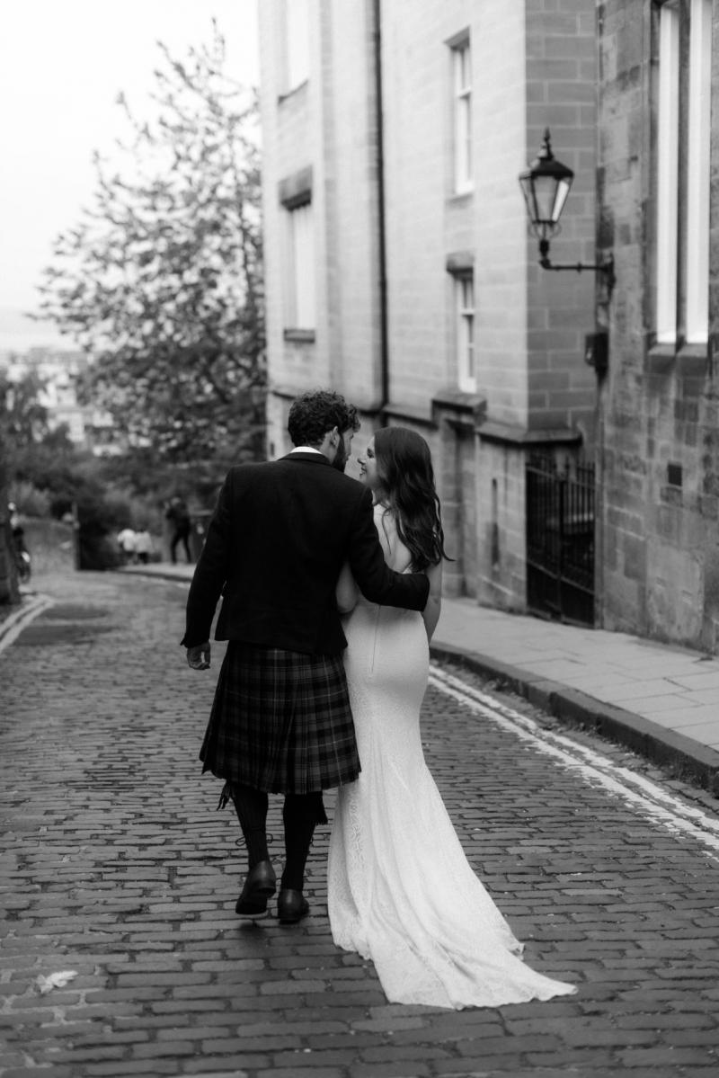 B&W photo of Mel and Robbie walking down a cobblestone street as her long lace train from her Jemma wedding dress trails behind.