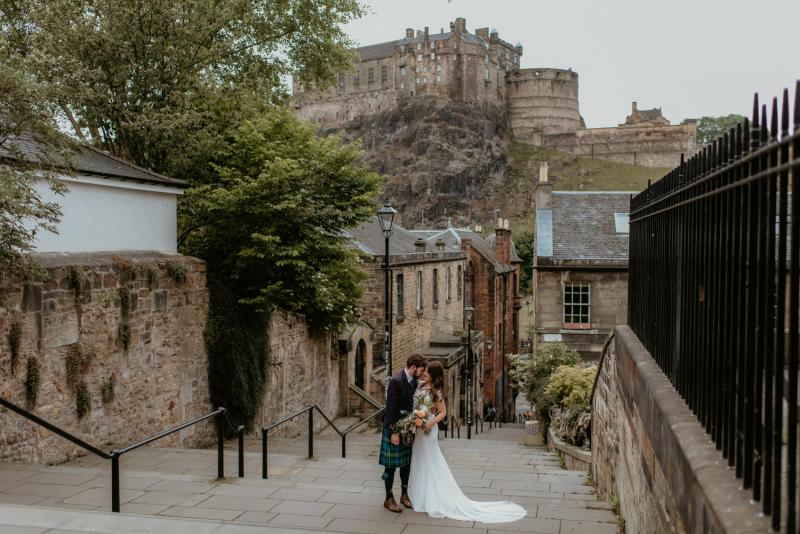 KWH real bride Mel and Robbie kiss on the old steps of Edinburgh. She wears the soft ivory Jemma wedding dress with a long lace train.