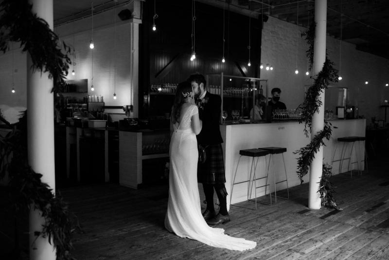 B&W photo of KWH real bride Mel and Robbie kissing at their reception bar. She wears the Jemma wedding dress which features a long lace train and low back.