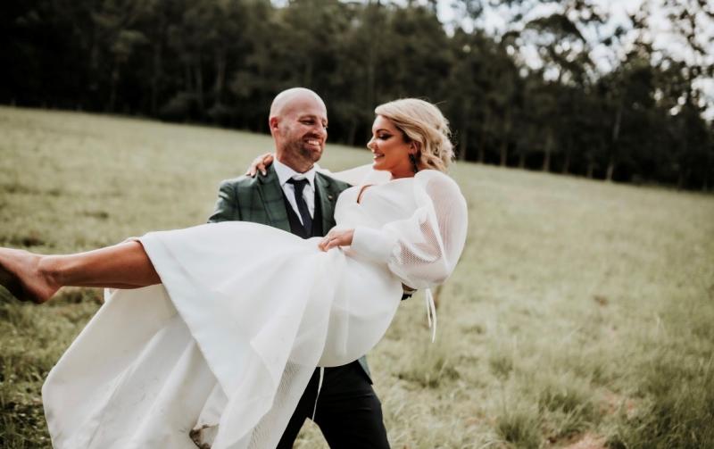 KWH real bride Caitlin being carried by Eoin in the grass. She wears the modern ivory Penelope wedding dress with fit and flare shape.