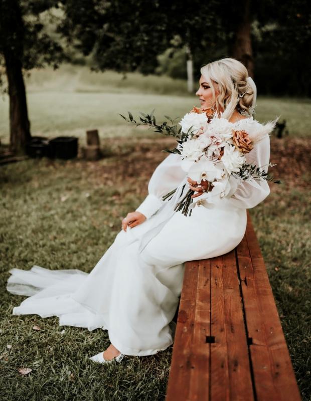KWH real bride Caitlin sitting on a wooden bench. She wears the long sleeve Penelope wedding dress