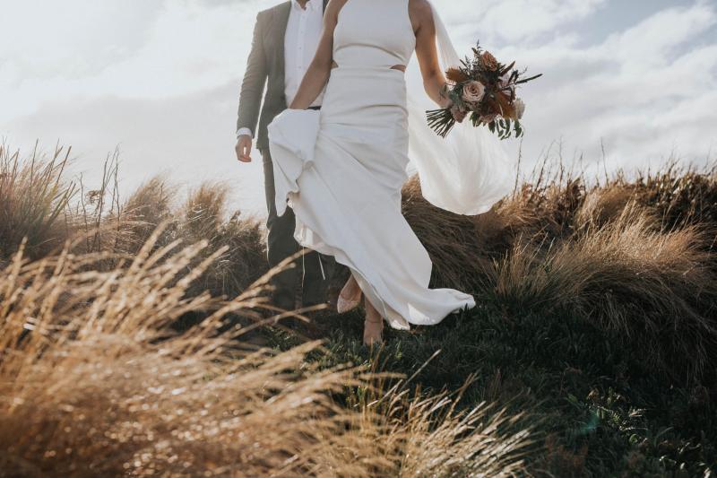 KWH real bride Ashleigh and Simon walk down the grassy hillside as she carries her native bridal bouquet and her Bridget dress' train in her hand.