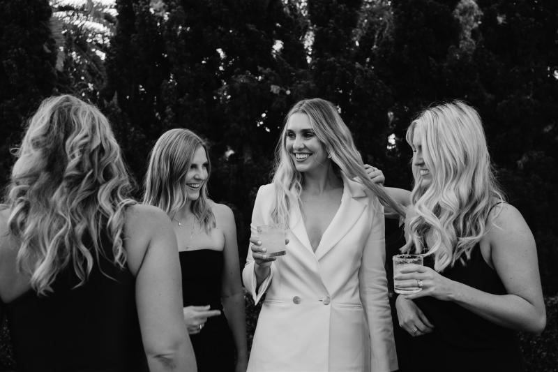 B&W image of KWH real bride Demi talking with her bridesmaids who wear black dresses. She wears the modern Charlie Danielle tailored bridal suit.