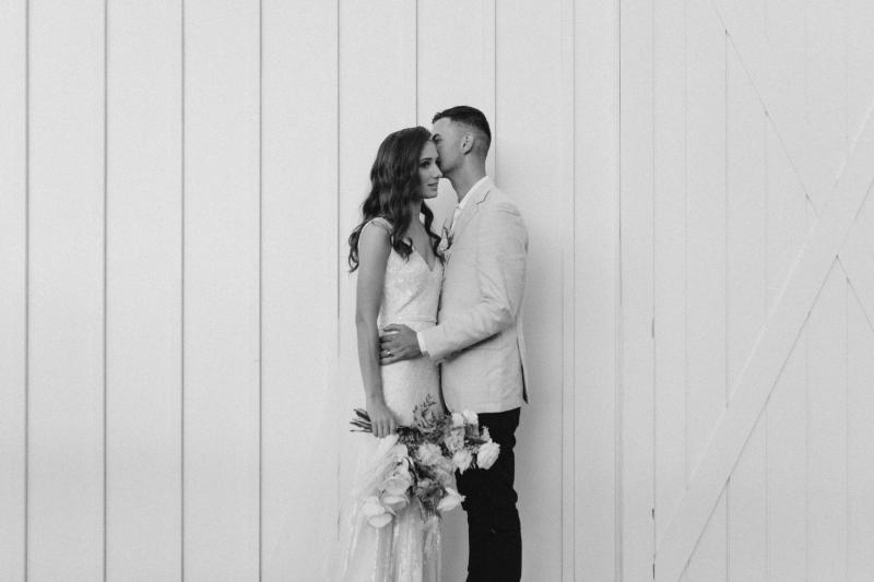 B&W image of KWH real bride Georgie and Mikey hugging in front of the white barn. She wears the Fontanne gown, a hand-beaded nontraditional wedding dress.