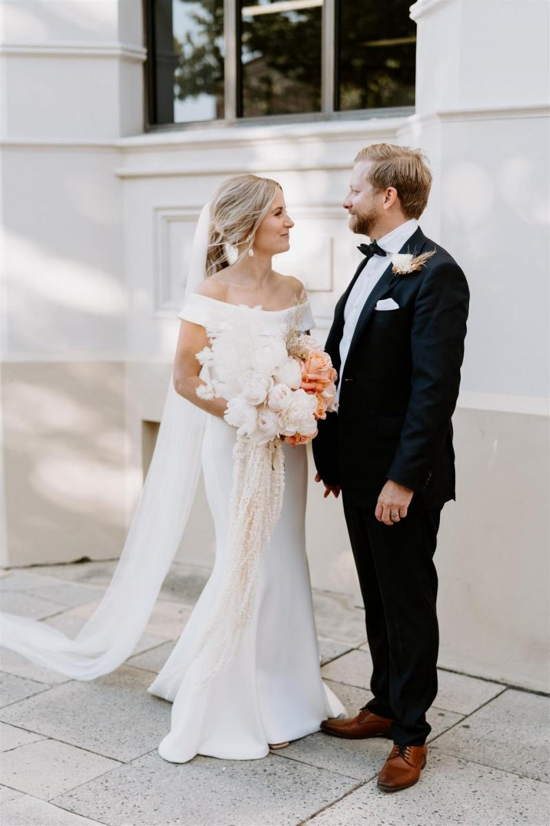 KWH real bride Shelley looks at her new husband Jack in front of a city building. She wears the simple Lauren wedding dress with trumpt train and fitted silhouette.