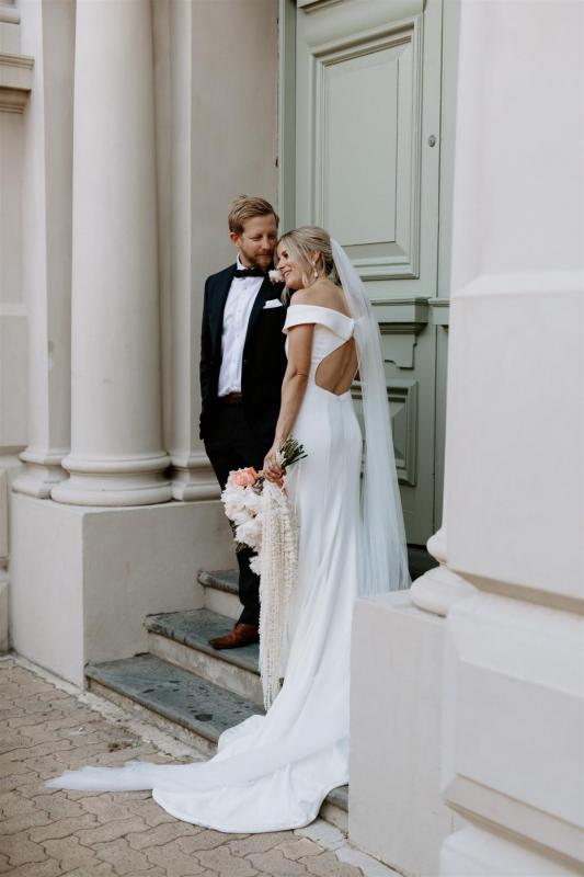 KWH real bride Shelley stands with Jack by a pale green door. She wears the sophisticated Lauren wedding dress with keyhole back and satin buttons.