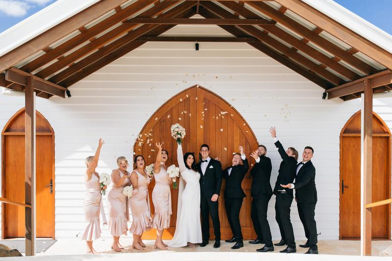 KWH real bride Rebekah and her bridal party celebrate out front the barn doors. She wears the ivory Nikki wedding dress with V-neck and long sleeves.