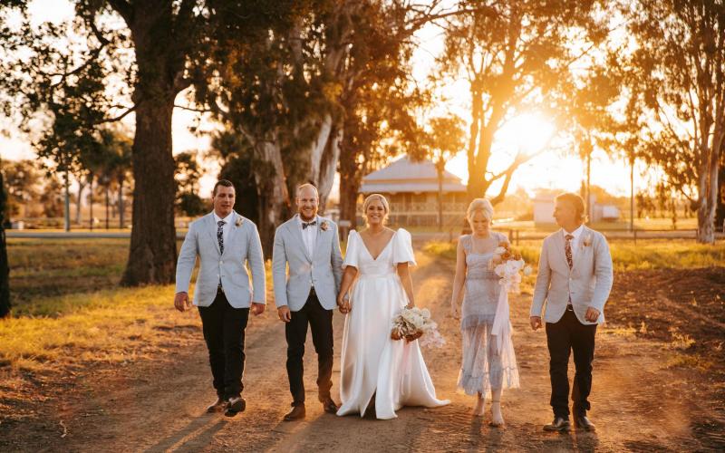 KWH real bride Izzy and Jake walk with their wedding party down a tree lined road. She wears the modern Taryn Camille wedding dress with over-sized sleeves.