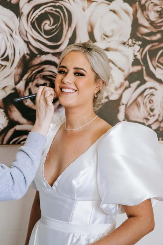 KWH real bride Izzie gets makeup applied to her face as she wears her ivory Taryn Camille wedding dress with large bubble sleeves.
