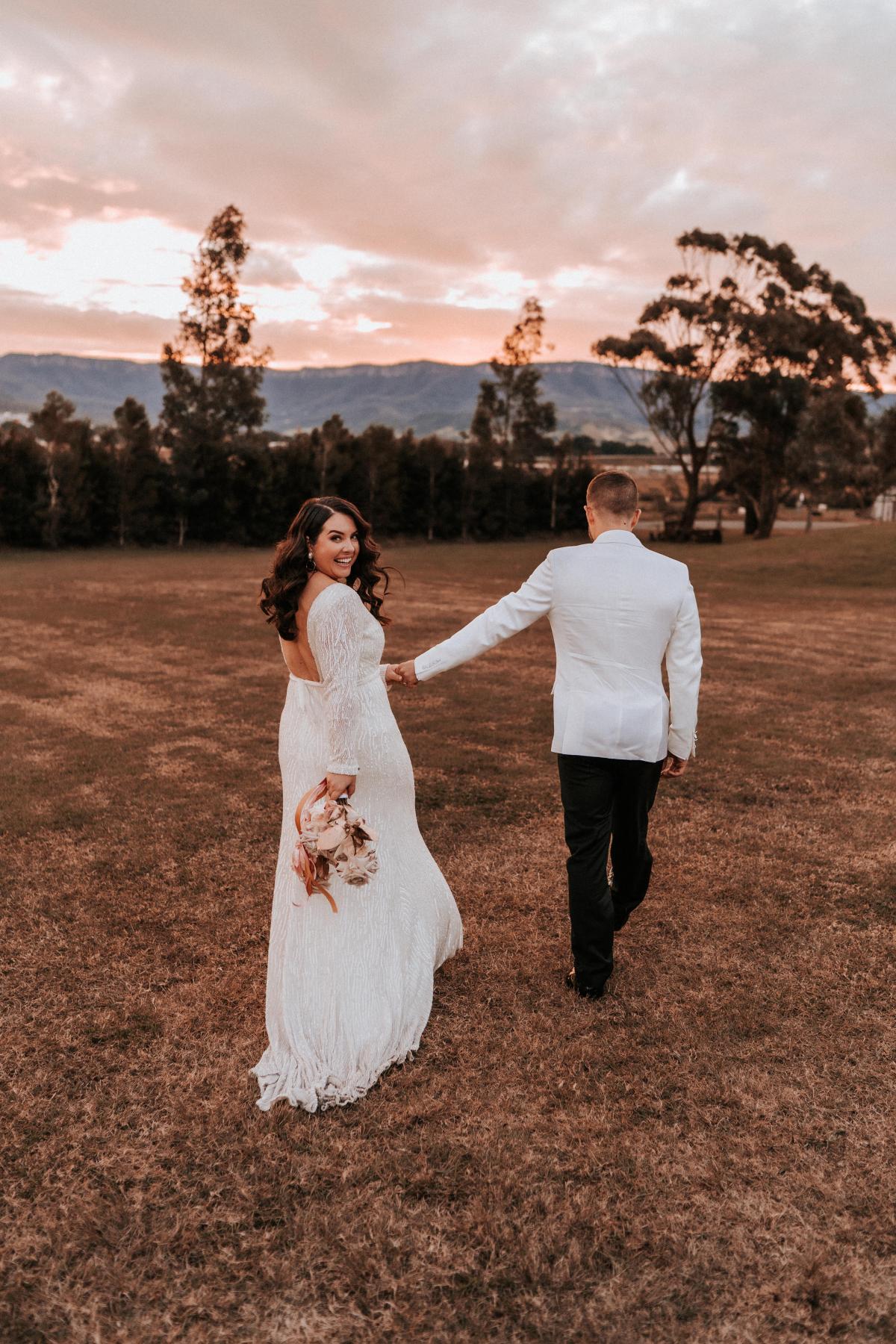 KWH real bride Katherine and Conor walk through a field together as the sunsets. She wears the Margareta gown, a backless high neck wedding dress