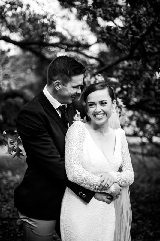 B&W image of KWH real bride Rachel and josh hugging after their vows. She wears the Rylie gown, a fit and flare lace wedding dress with long sleeves