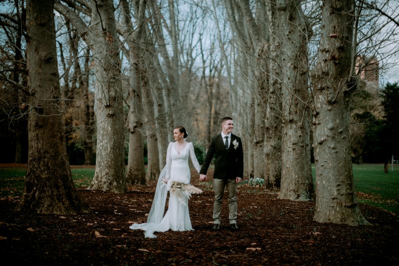 KWH real bride Rachel and Josh holding hands in the woods. She wears the Rylie gown, a fit and flare long sleeve vneck wedding dress