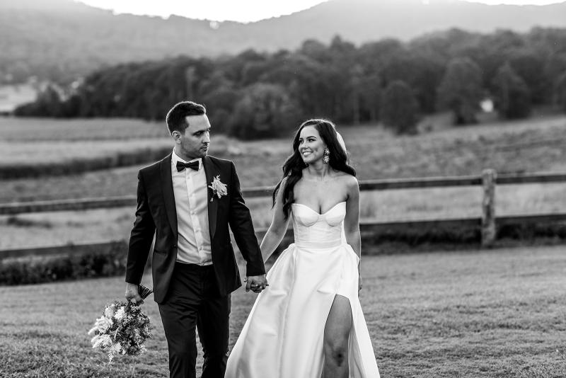B&W image of KWH real bride Hannah and Reece walking. She wears the Blake Camille gown, a modern strapless aline wedding dress with leg split.