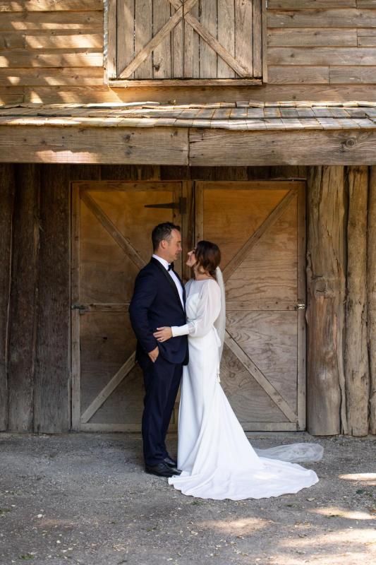 KWH real bride Erin and Stephen hug in front of barn doors. She wears the modern Brie gown, simple wedding dress with high neck and long sleeves.