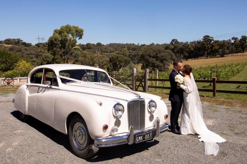 KWH real bride Brie stands with her new husband next to a classic car. She wears the modern Brie gown, simple wedding dress with high neck and long sleeves.
