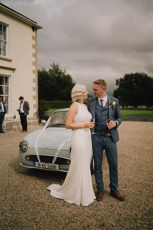 KWH real bride Laura and David stand in front of their vintage get away car as she wears the Cindy gown, a halter neck sequin wedding dress.