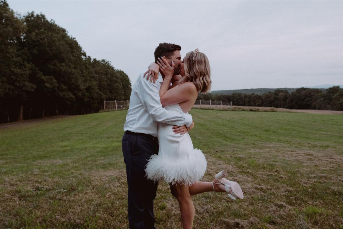 KWH real bride Kasey kisses brian in a field while wearing her Sherry mini gown, a simple short wedding dress with open back and feather detail.