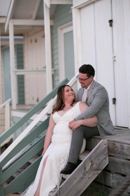 KWH real bride Meg and Tom sit on the beach house steps. She wears the effortless Bobby gown, a modern fit and flare wedding dress.